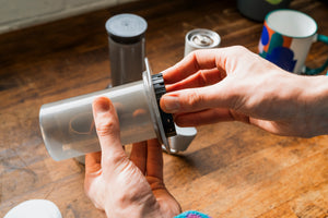 Close up photo of hands opening an AeroPress with coffee in the background