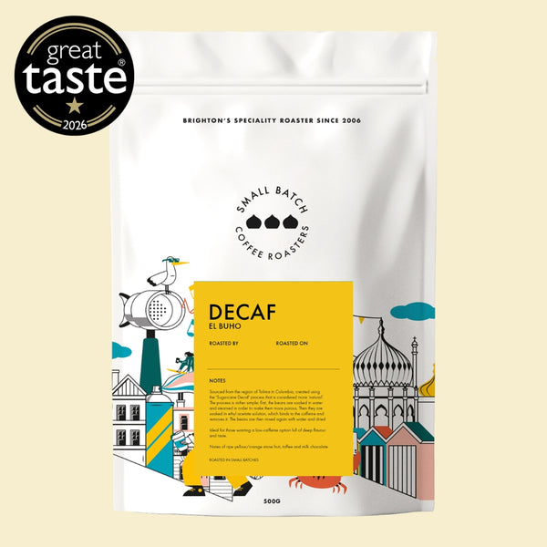 A stand-up coffee bag on a pale beige background with a black circular 'great taste 2026' award badge in the top-left corner. The package is white with a large yellow front label and colorful illustrated architectural graphics.