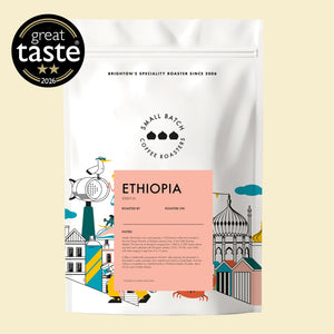 A white stand-up coffee bag labeled Small Batch Coffee Roasters Ethiopia, with a peach label panel and illustrated architectural and roasting-themed graphics. A black 'great taste 2025' badge appears in the top-left corner on a pale beige background.