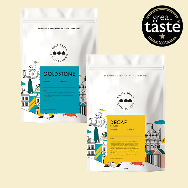 Two white coffee bean bags from Small Batch Coffee Roasters on a light beige background, one labeled Goldstone with a blue label and the other labeled Decaf with a yellow label. A black 'great taste 2026' badge is in the top right corner.