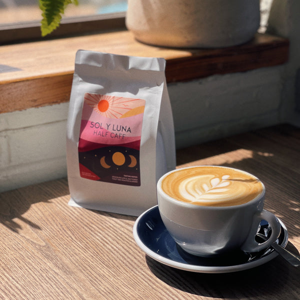 Cup of coffee with a bag of Sol y Luna coffee on a wooden surface with a plant in the background.
