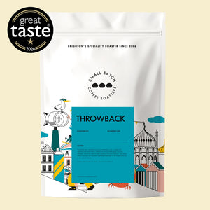 A white stand-up pouch of Brighton's Specialty Roaster Throwback coffee beans, featuring a bright blue label and colorful coastal illustrations, shown on a light beige background with a 'great taste 2026' badge in the top-left corner.