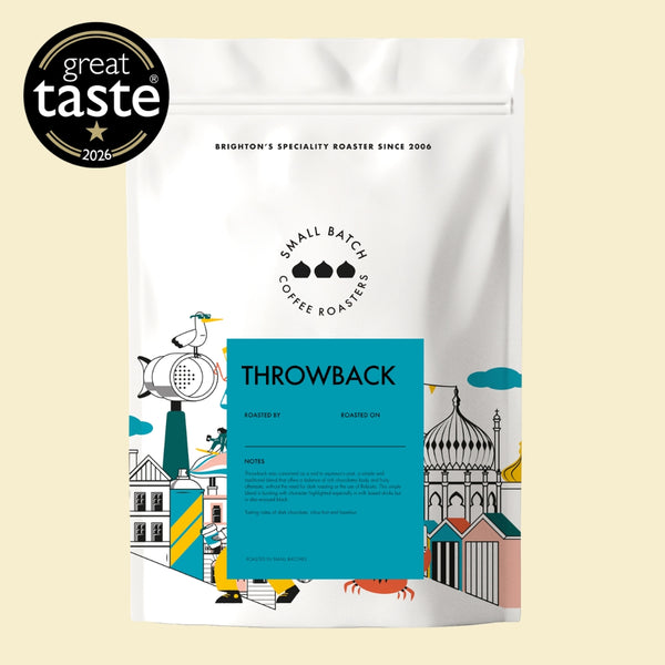 A white stand-up pouch of Brighton's Specialty Roaster Throwback coffee beans, featuring a bright blue label and colorful coastal illustrations, shown on a light beige background with a 'great taste 2026' badge in the top-left corner.